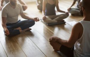 Menschen, die in einer Yogastunde mit gekreuzten Beinen und auf den Knien ruhenden Händen in einer Meditationshaltung auf einem Holzboden sitzen, verkörpern den Amema-Geist der Ruhe und Konzentration.