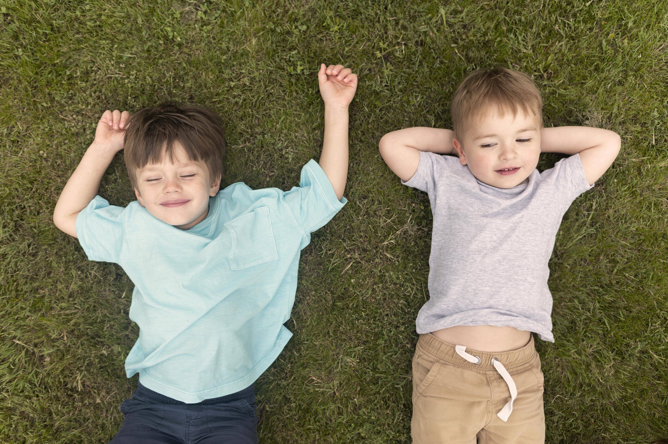 Zwei Kinder liegen entspannt auf dem Gras, eines mit einem blauen T-Shirt und das andere mit einem grauen T-Shirt.