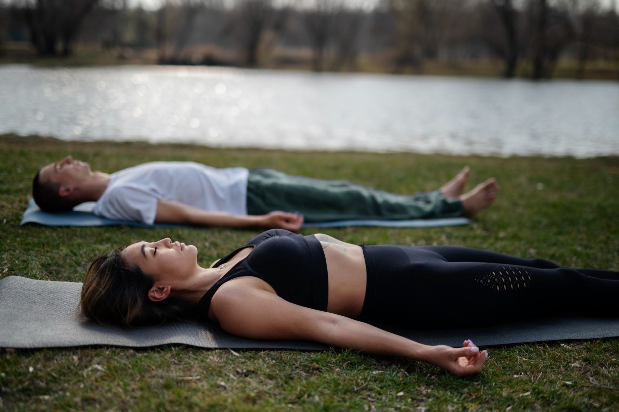 Zwei Menschen liegen auf Yogamatten auf einer Wiese in der Nähe eines Sees, die Augen geschlossen und die Arme an den Seiten, und üben „Transformatives Atmen“ sowie tiefe Entspannung oder Meditation im Freien.