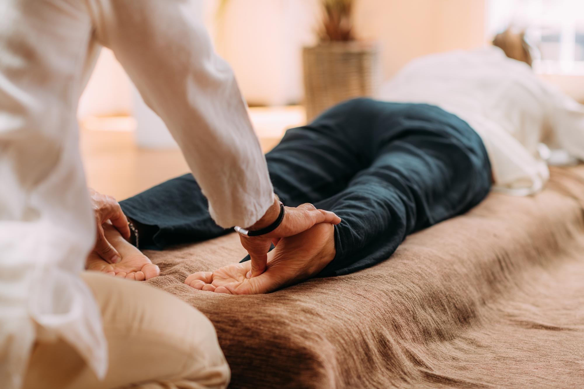 Eine Person liegt mit dem Gesicht nach unten auf einer Matte, während eine andere Person auf ihre Füße drückt und dabei möglicherweise eine therapeutische oder Massagetechnik wie Nuad Thai anwendet.