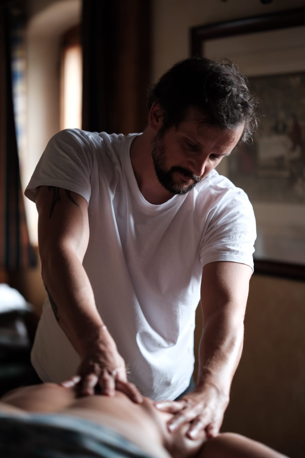 Ein Mann in einem weißen T-Shirt, möglicherweise ein erfahrener Masseur aus Tirol oder Atemtherapeut aus Tirol wie Lars Boob, gibt einer Person, die mit dem Gesicht nach unten in einem sanft beleuchteten Raum liegt, eine Rückenmassage.