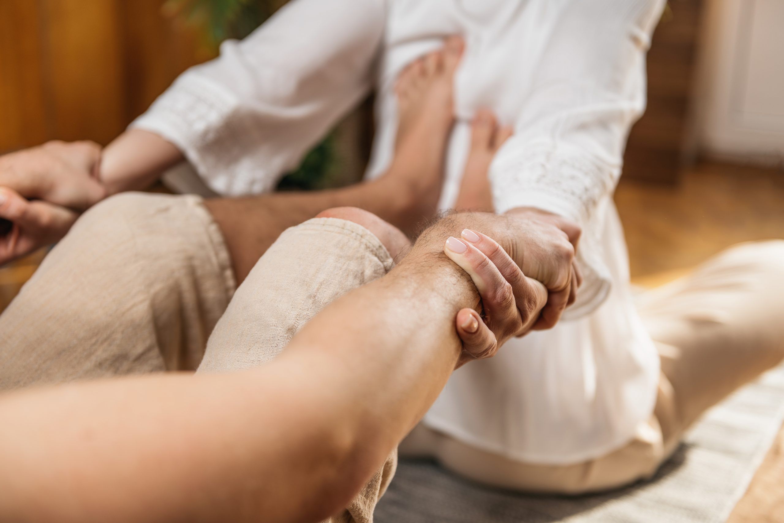 Eine Person unterstützt eine andere bei einer Dehnübung im Sitzen auf einer Matte, indem sie deren Arme festhält, während sich die andere Person mit ausgestreckten Beinen nach vorne beugt – eine von Nuad Thai inspirierte Technik.